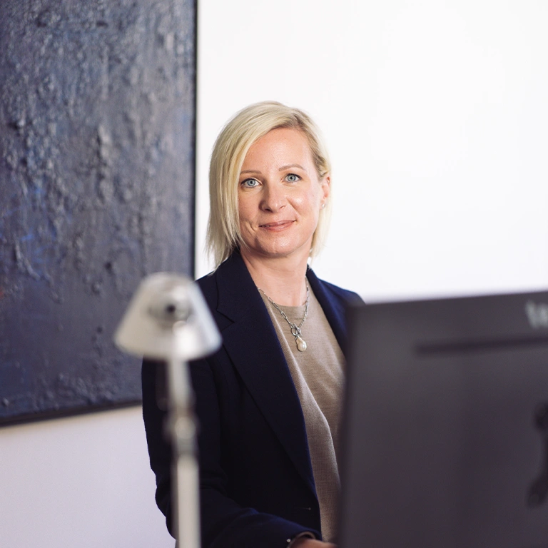 Professional woman with blonde hair wearing a navy blazer sits at a desk with a computer monitor, looking at the camera.